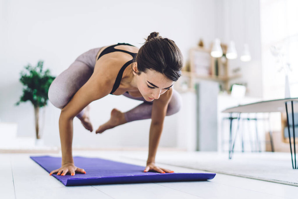 Full bode concentrated young woman with brown hair in sportswear doing handstand while practicing yoga on mat in modern studio in daytime - Foto, Imagen