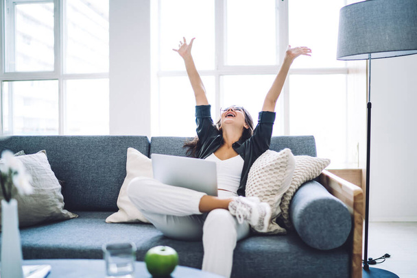 Emotional prosperous female blogger satisfied with successful project completing sitting with laptop computer, happy cheerful woman enjoying good news from social networks raising hands celebrating - 写真・画像