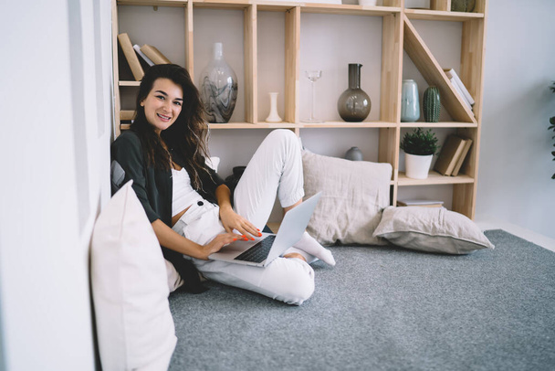 Side view of young female freelancer in casual clothes sitting on sofa and working on laptop while smiling and looking at camera - Foto, imagen
