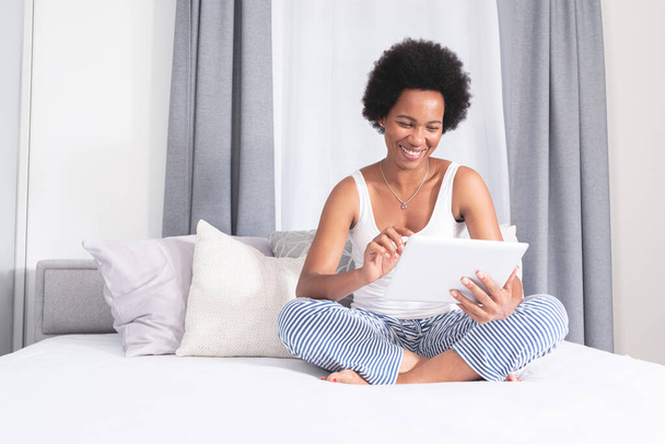 Happy african american woman using digital tablet sitting on bed at home - Fotó, kép