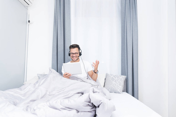 Happy man video chatting using digital tablet and headphones while relaxing on bed at home - Fotografie, Obrázek