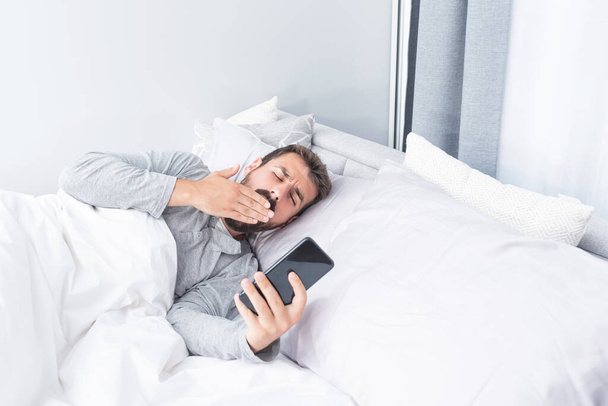 Young man yawning while using mobile phone lying in bed at home - Fotó, kép