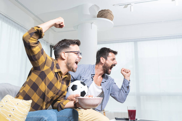 Twee jonge mannelijke vrienden schreeuwen terwijl ze voetbal kijken en popcorn eten. - Foto, afbeelding
