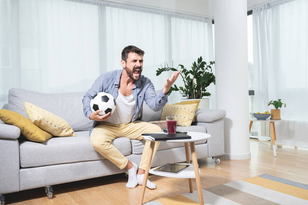 Разъяренный молодой футбольный фанат сидит на диване, держа мяч и крича во время просмотра матча - Фото, изображение