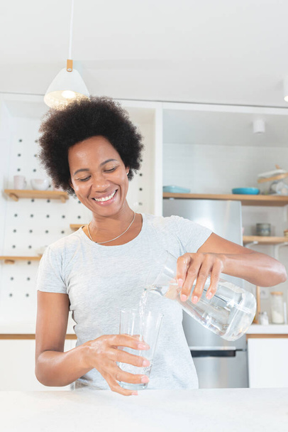 African American woman happily pouring water in drinking glass at kitchen - Foto, immagini
