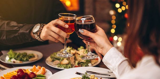 hands with glasses clinking against the background of Christmas tree lights and bonfires from a home fireplace over a table with delicious dishes - Photo, Image