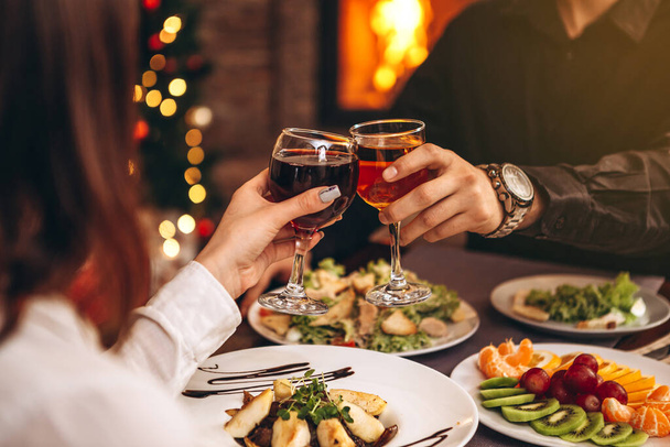 hands with glasses clinking against the background of Christmas tree lights and bonfires from a home fireplace over a table with delicious dishes - 写真・画像