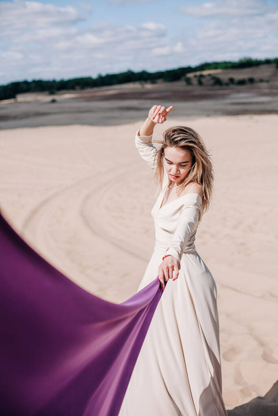 A young, slender girl in a beige dress with purple cloth in her hands posing in the desert in the wind - Fotografie, Obrázek