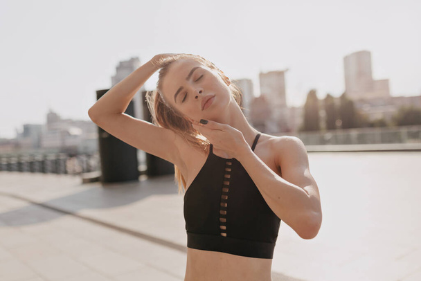 Foto de mujer joven fitness haciendo estiramiento al aire libre con los ojos cerrados y sonriendo. Una corredora en el campo por la mañana. Foto de alta calidad - Foto, Imagen