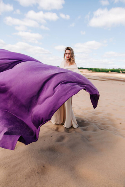 A young, slender girl in a beige dress with purple cloth in her hands posing in the desert in the wind - 写真・画像