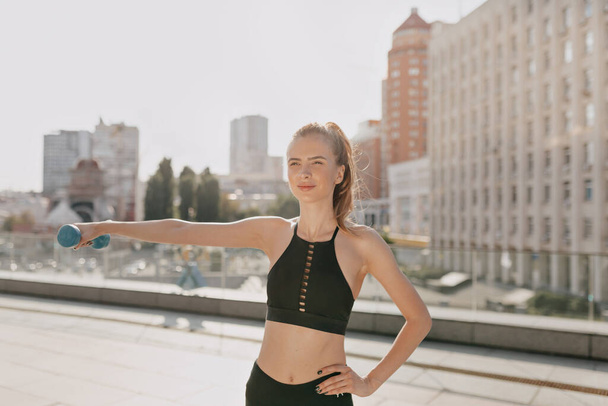 Happy exited woman in sportswear with hand dumbbells doing outside exercise in the city in sunny day . High quality photo - Foto, Bild