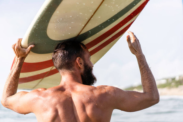 Mladý hipster muž s surfovací prkno na hlavě pozdravuje přátele s gestem - Guy baví dělat extrémní sport - sportovní vousatý muž trénink s surfem na moři - Životní styl, svoboda koncept - Fotografie, Obrázek