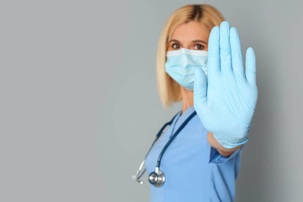 Doctor in protective mask showing stop gesture on grey background, space for text. Prevent spreading of coronavirus - Фото, изображение