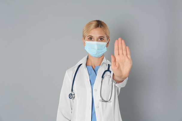 Doctor in protective mask showing stop gesture on grey background. Prevent spreading of coronavirus - Фото, зображення