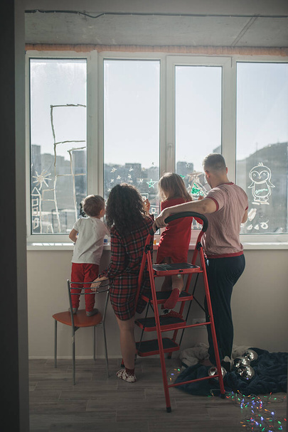 οικογένεια τεσσάρων παιδιών: μαμά, μπαμπάς, κόρη και γιος, ζωγραφίζουν με βούρτσες και μπογιές στο παράθυρο. Δραστηριότητες και ψυχαγωγία για όλη την οικογένεια κατά τη διάρκεια απομόνωσης και καραντίνας - Φωτογραφία, εικόνα