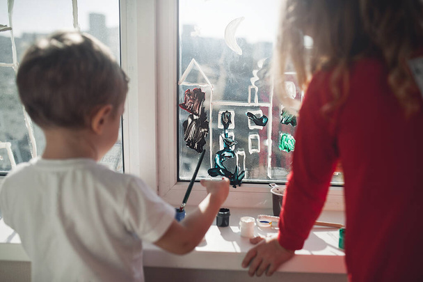 Schone kinderen, een jongen en een meisje tekenen met penselen en verf op het raam. Activiteiten en animatie voor kinderen tijdens isolatie en quarantaine - Foto, afbeelding