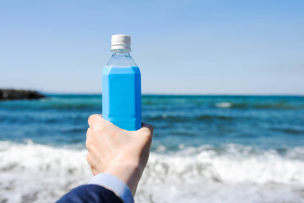 blauwe plastic fles in een licht gevilde hand tegen de achtergrond van een blauwe woedende zee met schuim. - Foto, afbeelding