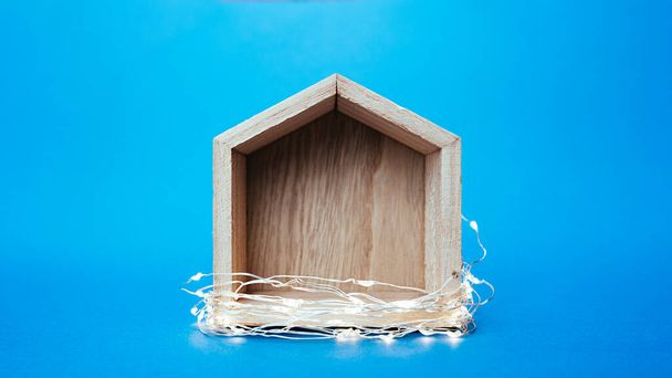 pequeña casa de juguete de madera gris envuelto guirnaldas y luces sobre un fondo azul brillante claro - Foto, imagen