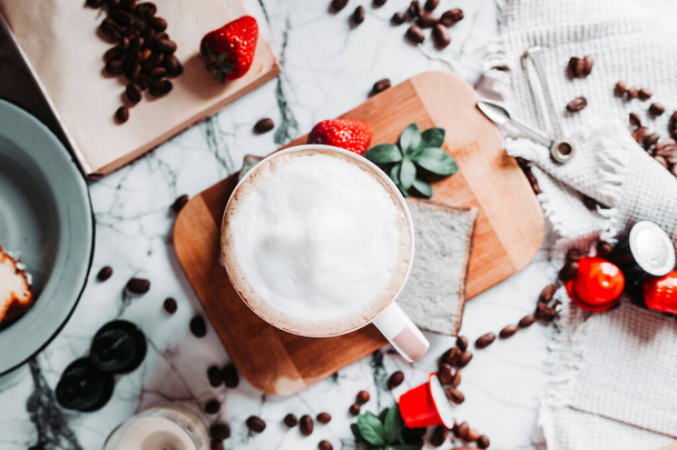 Leckere Kaffeepause trübt den Blick von oben. Eine volle Tasse Milchschaum auf einem Marmortisch mit einer türkischen Kaffeekanne, verstreuten Kaffeebohnen, Kaffeekapseln, Erdbeeren und Kerzen. - Foto, Bild