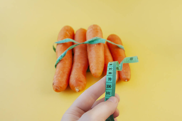 hand holding a centimeter long ribbon on a background of fresh orange peeled large carrots on a yellow background. weight control on quarantine and isolation - Photo, Image