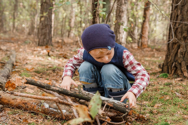 Ένα παιδί μαζεύει καυσόξυλα στο δάσος. Μικρέ ξυλοκόπε. Το αγόρι ψάχνει για παλιά κλαδιά δέντρων. Παιδί και καυσόξυλα. Φθινόπωρο. - Φωτογραφία, εικόνα