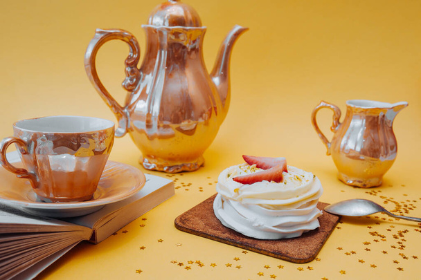 Belle collation lumineuse. Gâteau blanc à base de meringue, de crème et de fraises fraîches et d'ustensiles de thé doré : une tasse, une théière et un récipient pour crème sur fond jaune vif avec des étoiles dorées - Photo, image