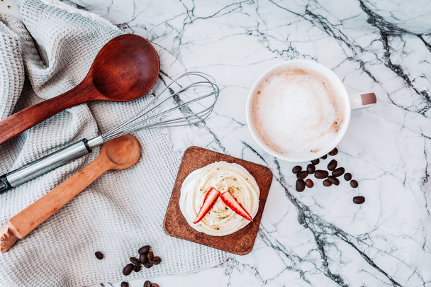 White dessert of meringue, cream and fresh strawberries, hot drink mug with whisk, wooden spoons and a waffle towel on white marble surface. Top view - Photo, Image