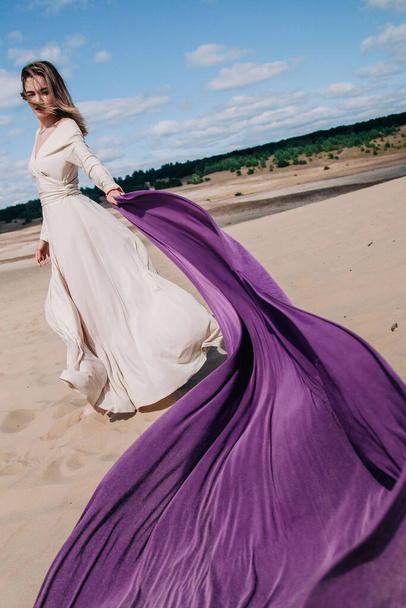 A young, slender girl in a beige dress with purple cloth in her hands posing in the desert in the wind - Foto, Imagen