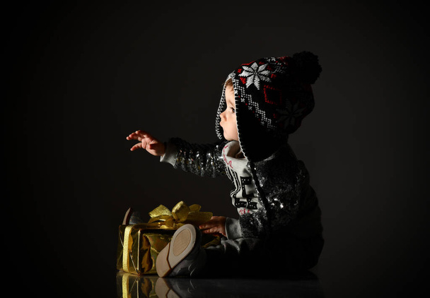 Kid in gray hat, suit and boots. She looking at snowflakes, holding golden gift box, sitting on floor. Black background. Close up - 写真・画像