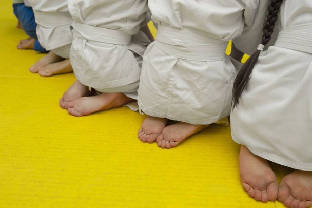 Nackte Beine und Absätze von Kindern in weißen Trainingsanzügen mit Wrestlinggurten auf einer gelben Matratze - Foto, Bild
