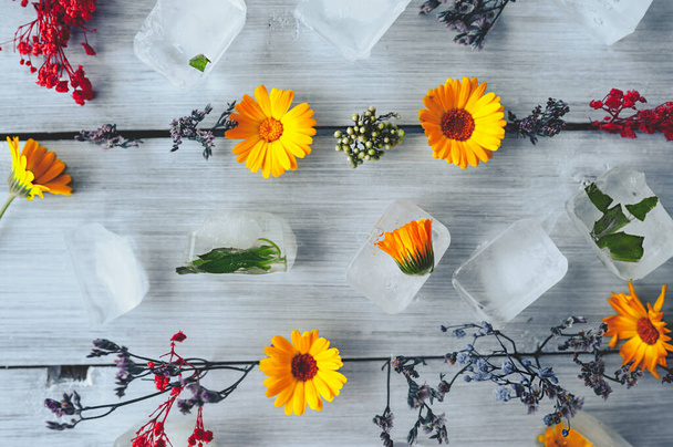 frozen ice cubes with orange calendula flower and green leaves and dry wild herbs on a light wooden surface, cosmetic layout - Photo, Image