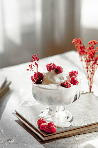 tál fehér fagylalt, piros bogyós lekvár, friss málna díszített piros virágok a világos asztalon a naplemente fény - Fotó, kép
