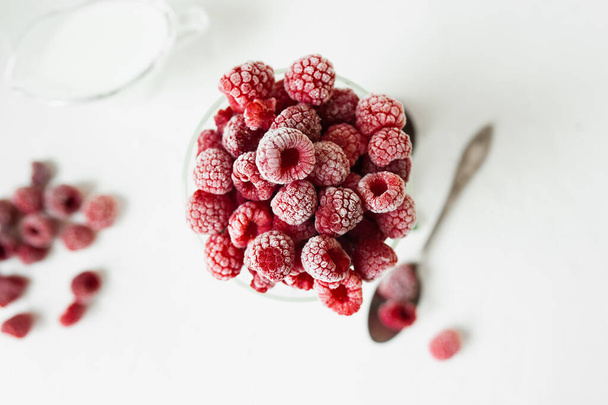 Свіжа заморожена червона красива малина у високій склянці на світлому столі зі срібною ложкою та розкиданими ягодами. Літнє меню, закуски в спекотну погоду. Сезонні стиглі ягоди
 - Фото, зображення