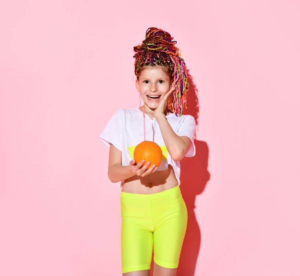 Nice little girl with multicolored cornrows on head laughing joyfully with closed eyes holding pineapple and orange in hands - Photo, Image