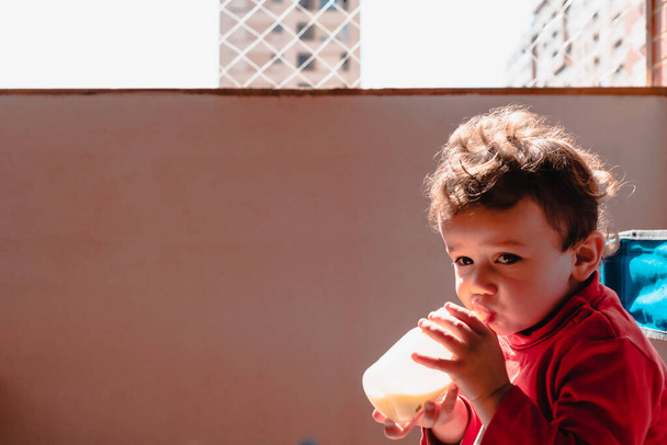Baby sitting sur le balcon de sa maison au soleil boit le lait d'une bouteille par elle-même. - Photo, image