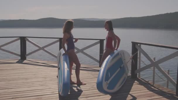 Full shot of young women in swimsuits standing on lake pier leaning on sup boards chatting - Footage, Video