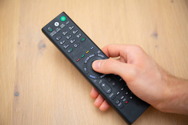 TV remote control in a male hand, with finger on a navigation button, on wooden background. Pressing a button on a remote control to turn on a television, top view, close-up. - Fotoğraf, Görsel