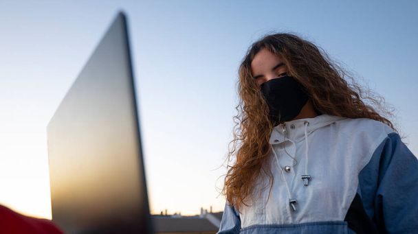 Brunette student with long curly hair wearing winter clothes on the roof of a flat with laptop doing homework and wearing a mask in times of a coronavirus pandemic. - Foto, Bild