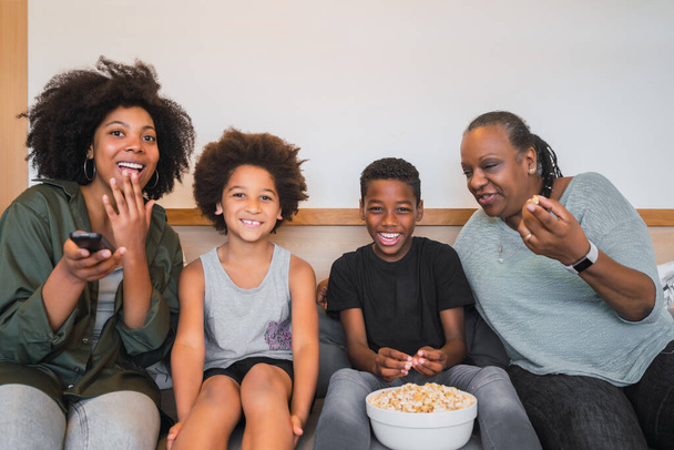 Portrait of African American grandmother, mother and children watching a movie and eating popcorn while sitting on sofa at home. Family and lifestyle concept. - Fotó, kép
