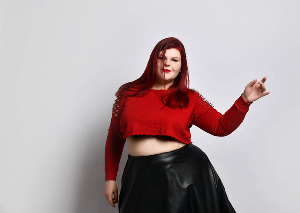 Obese ginger lady in red spiked top, black bra and leather skirt. She is dancing, posing isolated on white photo background - Photo, image