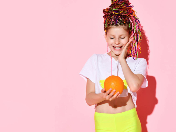 Nice little girl with multicolored cornrows on head laughing joyfully with closed eyes holding pineapple and orange in hands - Zdjęcie, obraz