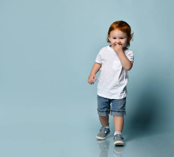 Little ginger toddler boy or girl in white t-shirt, socks and shoes, denim shorts. Child is smiling, walking on blue background - Zdjęcie, obraz