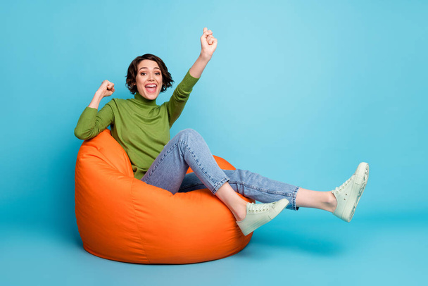 Full length photo of excited girl sit bean chair raise fists celebrate lucky lottery win wear jumper isolated blue color background - Foto, imagen