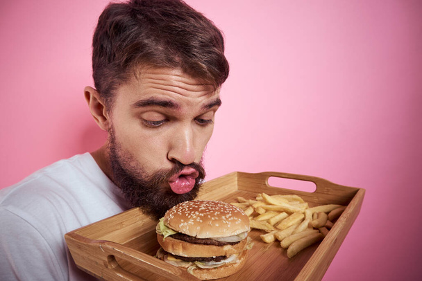 Mężczyzna z tacą jedzenia w ręku hamburger frytki i fast food kalorii różowy tło portret zbliżenie - Zdjęcie, obraz
