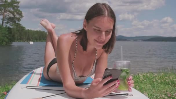 Gros plan moyen de la femme moderne avec téléphone portable dans les mains défilant alimentation et sirotant boisson rafraîchissante couché sur le bord du lac - Séquence, vidéo