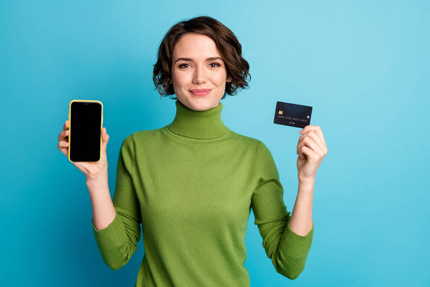 Portrait of positive girl hold smartphone credit card enjoy online banking paying modern technology wear green style stylish trendy jumper isolated blue color background - 写真・画像