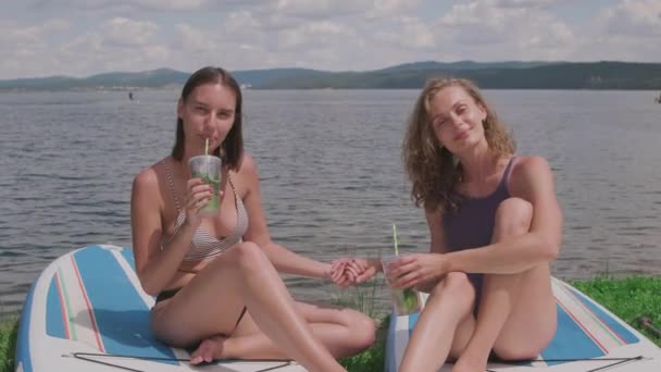 Mittlere Aufnahme von jungen, gut gelaunten Frauen, die Cocktails am See schlürfen und dabei fröhlich in die Kamera lächeln - Filmmaterial, Video