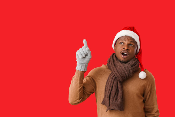 Uomo afro-americano in cappello di Babbo Natale e guanti che puntano a qualcosa sullo sfondo di colore - Foto, immagini