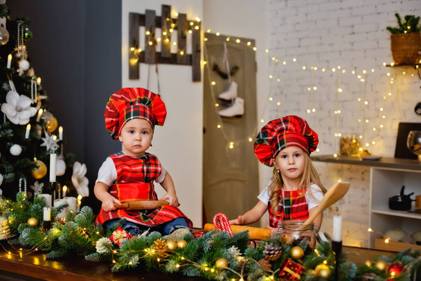 Ένα παιδί με καπέλο σεφ και ποδιά ετοιμάζει ζύμη μπισκότων για τα Χριστούγεννα. Τα παιδιά ετοιμάζουν γλυκά στην κουζίνα με χριστουγεννιάτικο δέντρο σε μια χιονισμένη χειμωνιάτικη μέρα. - Φωτογραφία, εικόνα