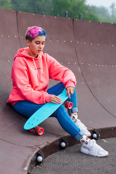 Stylish young woman with short colored hair sitting with her blue plastic skateboard in skatepark. Youth concept. - Photo, Image